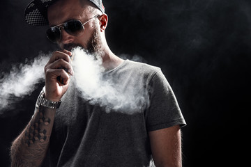 Pensive Black bearded male dressed in a grey shirt, sunglasses and baseball cap vaping. man in...