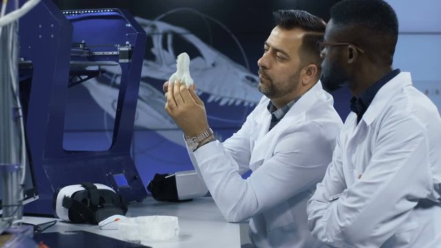 Multiracial men in white apparels sitting at table in laboratory and discussing 3-D model of dinosaur skull.