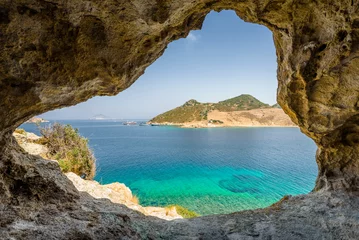 Fototapeten Beautiful sunny view through a rocky cave to the greek blue sea with crystal clear water from an a hill with boats cruising fishing surrounded by mountains, Patmos Island, Kos, Dodecanese/ Greece  © Thomas Jastram