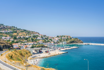 Fototapeta premium Beautiful sunny greek village town white house and harbor view to the aegean blue sea with crystal clear water and fishing boats cruising yacht white houses, Ikaria Island, Evdilos, Sporade, Greece