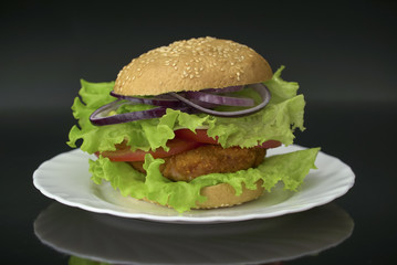 Tasty burger on white plate against black background. Homemade hamburger with chicken cutlet, tomatoes, onion and salad.