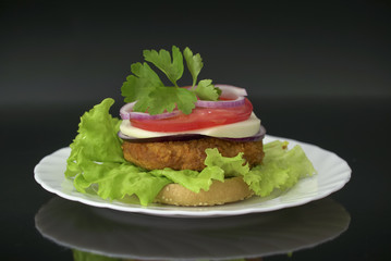 Tasty burger on white plate against black background. Homemade hamburger with fresh vegetables, cheese and chicken cutlet.