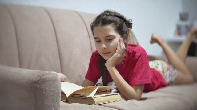 girl teen reading a book lying on sofa indoors. girl kid reading a book lying indoors on a sofa