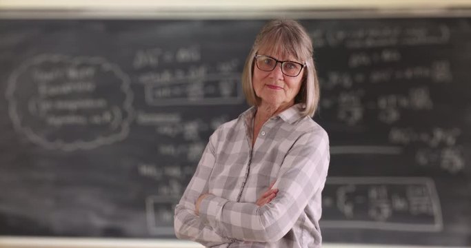 Confident senior woman teacher with arms crossed standing  in front of chalkboard, Mature lady looking proud crossing arms inside classroom, 4k