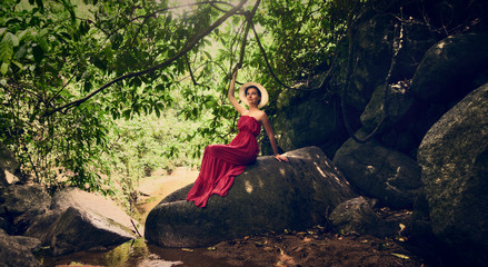 A girl in a hat sits on a rock in the jungle. Girl in a red dress.