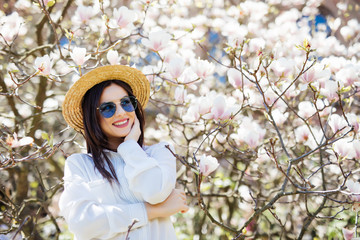 Beauty young woman in sunglasses and straw hat standing near Magnolia blossoming flowers tree in spring graden