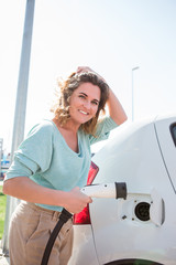 A woman stands at the charging station and holds a plug of the charger for an electric car
