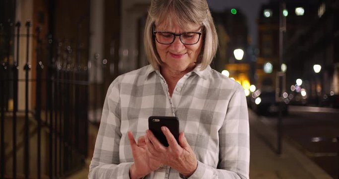 Charming old lady using portable technology to communicate outside in city at night, Smiling senior woman using phone to send text message on city sidewalk at night, 4k