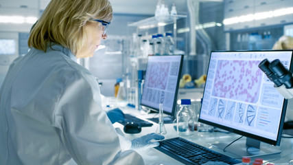 Senior Female Scientist Works On Computer in a Modern Laboratory. Her Colleagues are Working Beside...