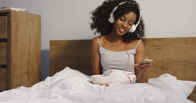 Young pretty African American curly woman in the headphones sitting on the unmade bed in the morning, listening to the music and eating muesli for the breakfast. Indoor