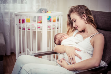Breastfeeding baby near crib. Pretty mother holding her newborn child. Mom nursing baby. Beautiful woman and new born relax at home in nursery interior. Blondе mother breast feeding baby.