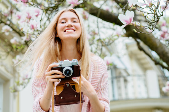 Positive blond woman traveler take photos by camera under magnolia blossoms tree. Female use retro camera under sakura in full blooming.