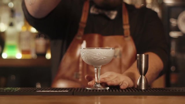 Barman is pouring white cocktail in a goblet in a bar of restaurant. He is putting paper decor on an edge of glass and moving it forward