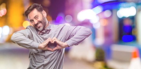 Middle age man, with beard and bow tie happy showing love with hands in heart shape expressing healthy and marriage symbol at night club