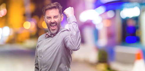 Middle age man, with beard and bow tie happy and excited expressing winning gesture. Successful and celebrating victory, triumphant at night club