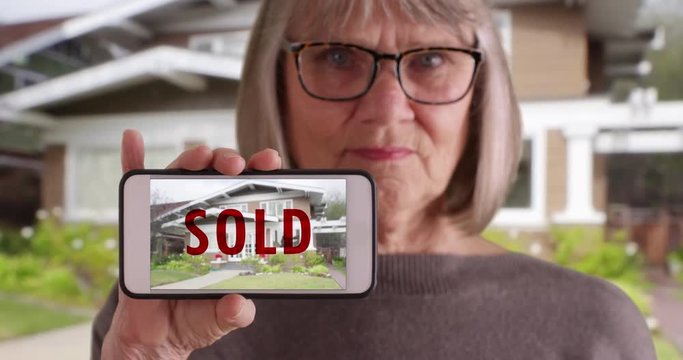 Elderly white woman holding phone with picture of home that was recently sold, Serious looking old woman showing picture of sold house on smartphone to camera, 4k