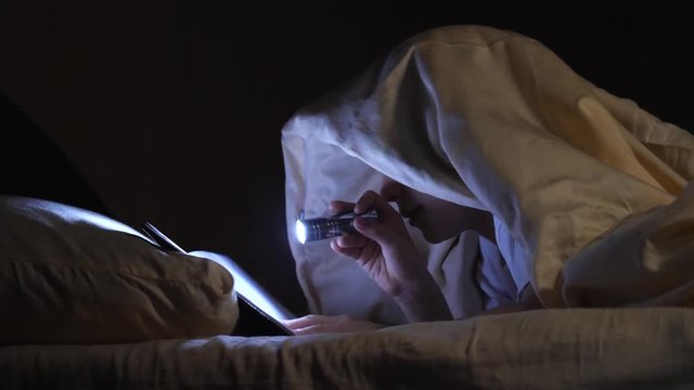 A child reads a book under blankets with a flashlight at night. Enthusiastic boy