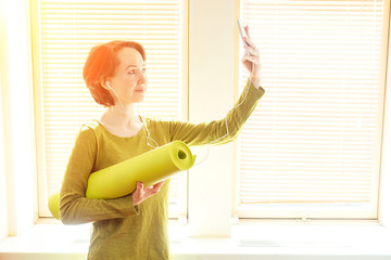 cheerful brunette woman in a green blouse holds a roll of mate and makes selfie on the phone near the window in a yoga class. smartphone in the hands of a yoga coach