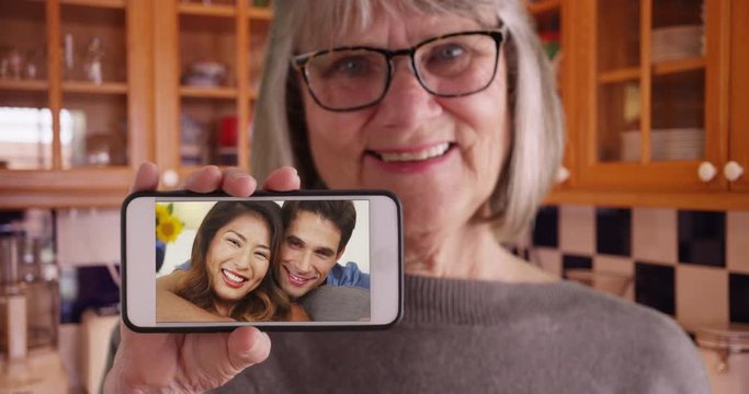Old lady holding phone to camera while video chatting with son and his girlfriend, White elderly female holding phone with happy white man and Asian woman on the screen, 4k