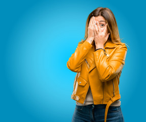 Beautiful young woman smiling having shy look peeking through her fingers, covering face with hands looking confusedly broadly, blue background