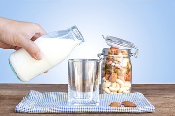 Woman pouring milk into a glass and a bottle of dried fruit, various nuts on the wooden table.