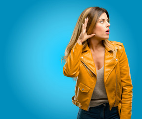 Beautiful young woman holding hand near ear trying to listen to interesting news expressing communication concept and gossip, blue background