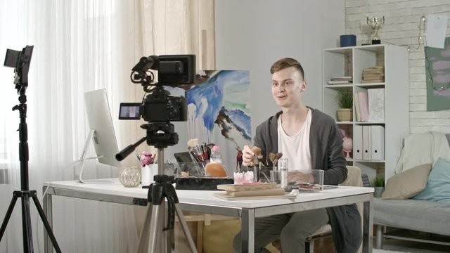 PAN of cheerful male teenage beauty blogger shooting makeup tutorial: he sitting before camera on tripod and waving hello, then explaining what brand of micellar water he using