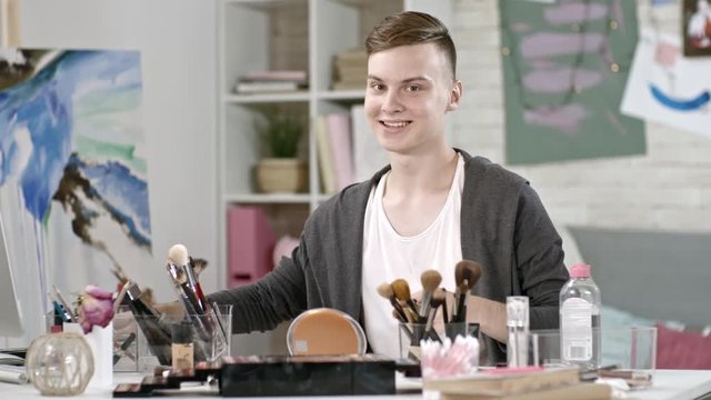 Medium shot of cheerful teenage boy with stylish haircut using clapperboard and looking at camera while showing makeup brushes