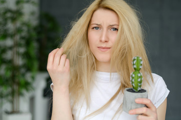 Naklejka premium Damaged Hair. Beautiful Sad Young Woman With Long Disheveled Hair. Closeup Portrait Of Female Model Holding Messy Unbrushed Dry Hair In Hands. Hair Damage, Health And Beauty Concept With Cactus