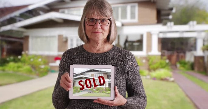 Senior Caucasian female holding tablet with picture of recently sold home, Casual looking old woman with pad device showing sold house outside residential home, 4k