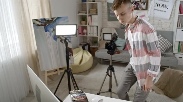 Tilt down of male teenage beauty blogger putting down video camera on tripod and sitting down to work with footage on computer