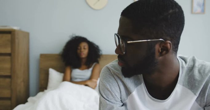 Close up of the upset young African American man sitting on the bed, turning his head and looking at his beautiful wife or girlfriend who sitting in the bed on the blured background. Indoors
