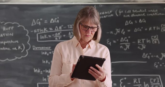 Independent senior woman teacher working on tablet device in front of chalkboard, Mature lady enjoying portable tech in classroom setting in front of blackboard, 4k
