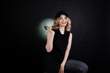 Studio portrait of blonde girl in black wear and cap with mobile phone at hand against dark background.
