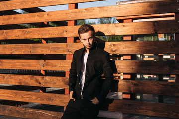 The guy in stylish clothes leans on a wooden fence and looks at the camera lens. sun rays shine on the guy