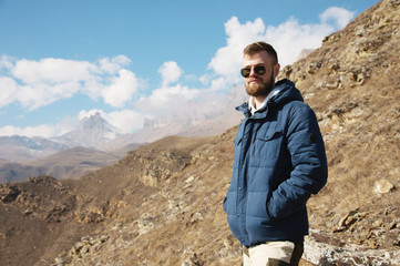 Hipster traveler in a down jacket and sunglasses stands on a mountain slope against the backdrop of epic rocks and smiles. The concept of finding happiness in the mountains
