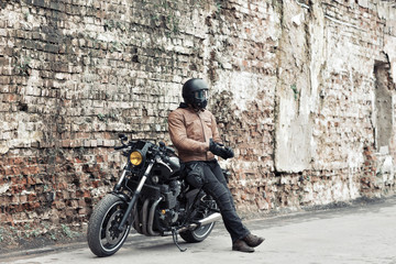 Obraz na płótnie Canvas Man a motorcyclist standing with helmet near her bike, brick wall of garage background. Male in a leather jacket and tight pants
