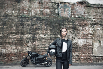 Plakat Woman a motorcyclist standing with helmet in hand near her bike, brick wall of garage background. Girl in a leather jacket and tight pants, holding motorcycle helmet in hands.