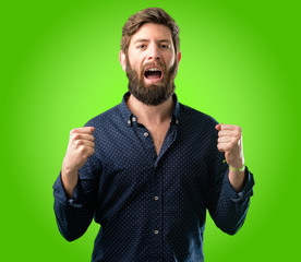 Young hipster man with big beard happy and excited expressing winning gesture. Successful and celebrating victory, triumphant over green background