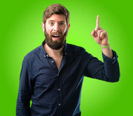 Young hipster man with big beard happy and surprised cheering expressing wow gesture pointing up over green background