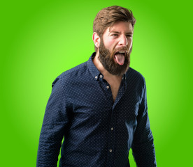 Obraz premium Young hipster man with big beard sticking out tongue at camera at sign of disobedience, protest and disrespect over green background