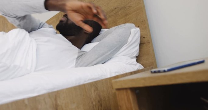 Close Up Of The Young African American Attractive Man Waking Up In The Morning In His Bed, Looking At The Time On The Smartphone And Keeping On Sleeping. Turning Off The Larm Ringing. Indoors