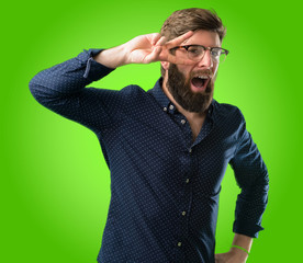 Naklejka premium Young hipster man with big beard looking at camera through her fingers over green background