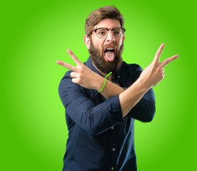 Young hipster man with big beard looking at camera showing tong and making victory sign with fingers over green background