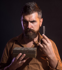 Brutal caucasian hipster, serious bearded man with long beard holds in hands dangerous razor,...