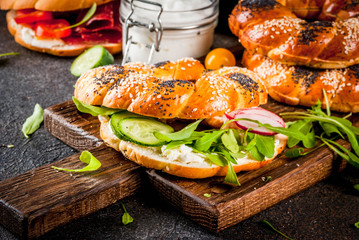 Variety of homemade bagels sandwiches with sesame and poppy seeds, cream cheese,  ham, radish, arugula, cherry tomatoes, cucumbers, with ingredients on dark concrete background