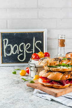 Variety of homemade bagels sandwiches with sesame and poppy seeds, cream cheese,  ham, radish, arugula, cherry tomatoes, cucumbers, white grey textured background copy space