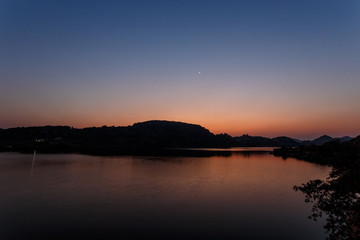 Fototapeta na wymiar Amazing twilight. The lake is waiting for nightfall. The sun has set by the mountain. Multicolored sky. Silhouettes of mountains. The first stars are lit. New moon.India. Hampi. Magical lake.