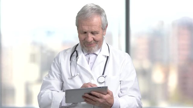Cheerful senior doctor using digital tablet. Elderly male doctor in white coat texting a message on blurred background.