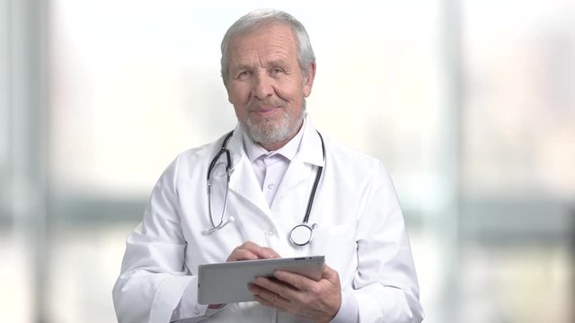 Smiling elderly doctor with pc tablet. Cheerful caucasian doctor using computer tablet on abstract blurred background.